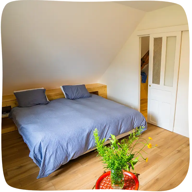 Chambre 1 - Lit double avec des draps bleu clairs. On voit la porte d'entrée au milieu à droite. On voit un petit tabouret rouge en avant plan, devant le lit, avec un bouquet de fleurs verte.