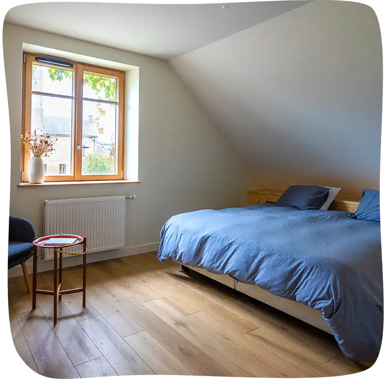 Chambre 1 - Lit double avec des draps bleus clairs. on voit la fenêtre sur le mur en face à gauche. On voit un petit tabouret rouge et un fauteul 1 place juste devant ce tabouret dans le coin à gauche