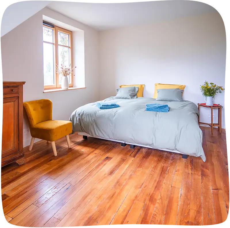 Chambre 2 – Lit double avec des draps bleu tournant vers le gris clair avec 4 coussins dont 2 de la même couleur et deux jaunes. Il y a une table de nuit du côté gauche du lit avec un bouquet de fleurs dessus. On voit également un fauteuil moutarde sans accoudoirs devant le lit et le début d'une armoire en bois. Le sol est un parquet en bois.