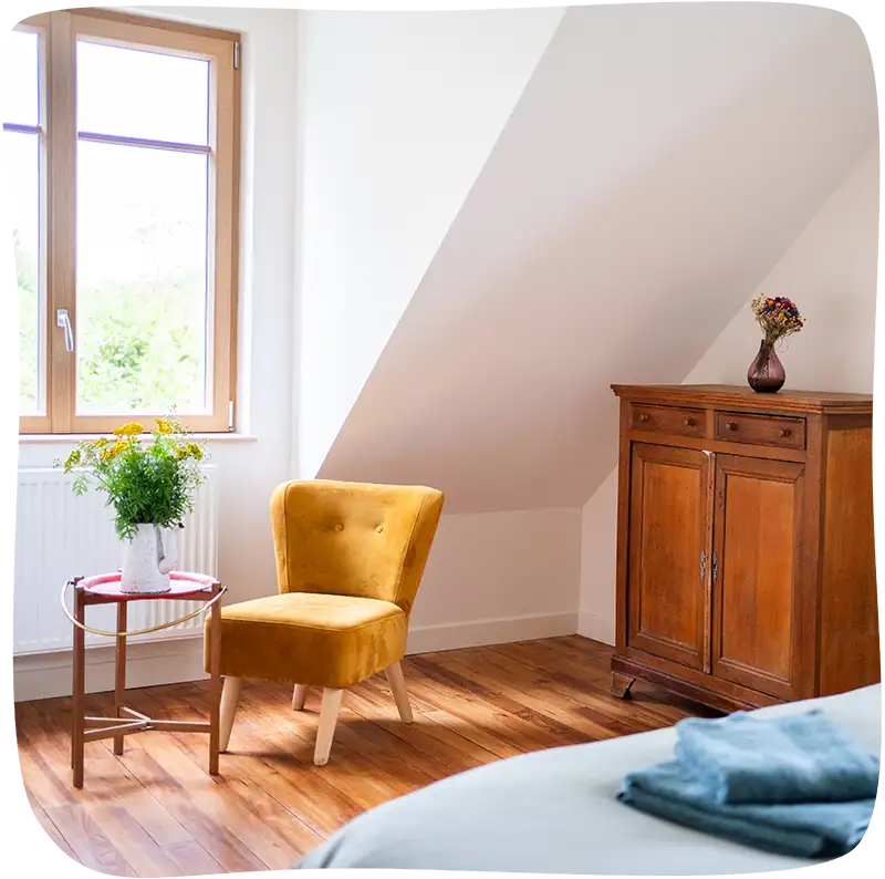 Chambre 2 – On voit le bout du lit avec des draps bleu tournant vers le gris clair en premier plan et le fauteuil moutarde ainsi que l'armoire en bois et un tabouret rouge avec un pot de fleurs jaunes et vertes eu milieu du plan. Le fauteuil se trouve juste devant une grande fenêtre lumineuse