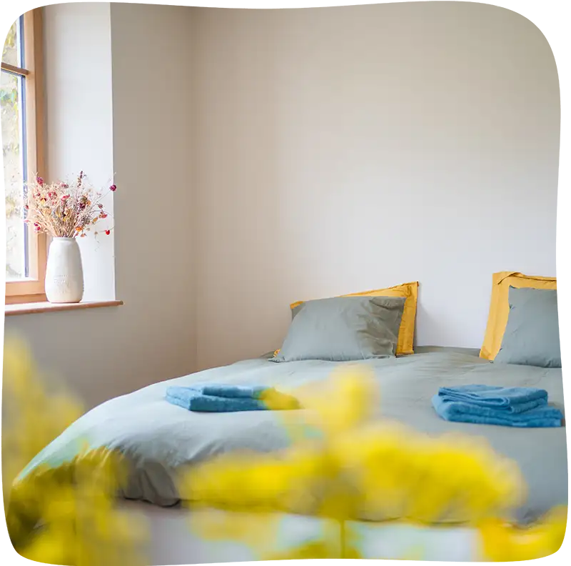 Chambre 2 – Vue proche du lit avec des draps bleu tournant vers le gris clair avec 4 coussins dont 2 de la même couleur et deux jaunes. On peut aperçevoir un bout d'une fenêtre lumineuse. Il y a des essuies de bain bleus sur le lit. Le premier plan est un flou de fleurs au bout jaune