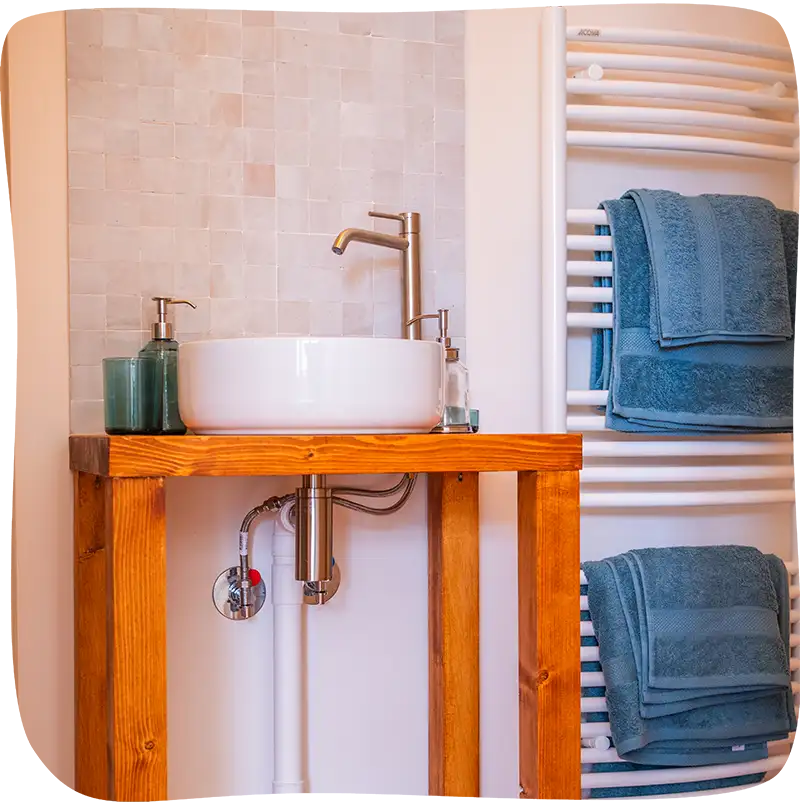 Chambre 2 – Evier de la salle de bain sur un meuble en bois. On y retrouve un savon main et un verre. Il y a un porte serviette mural chauffant à droite avec 2 paires d'essuies bleu