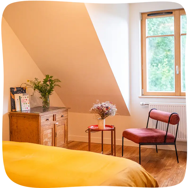 Chambre 3 – On voit le bout du lit avec des draps jaunes moutarde en premier plan et un fauteuil rouge ainsi qu'une armoire en bois et un tabouret rouge avec un pot de fleurs jaunes et vertes eu milieu du plan. Le fauteuil se trouve juste devant une grande fenêtre lumineuse