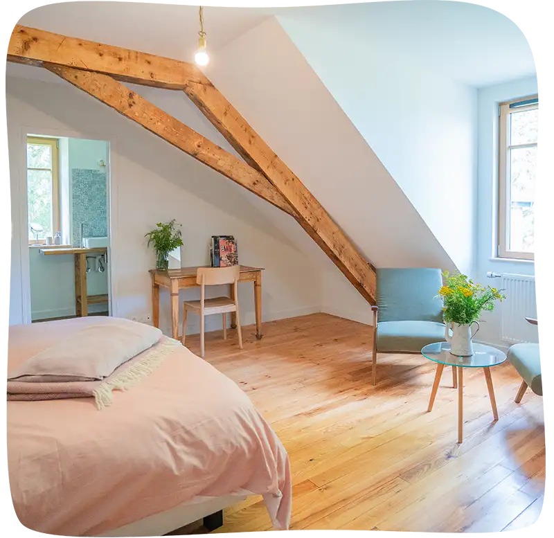 Bout de lit avec des draps rose pale et un plaid sur la house de couette. Il y a un bureau dans le fond à gauche avec une chaise en bois rangée. Il y a un pot de fleurs et des livres sur le bureau. On apercoit l'évier de la salle de bain à travers la porte ouverte à gauche du bureau en bois. On voit un 2 fauteuils et une table basse en verre a droite. Ces fauteuils sont devant une grande fenêtre lumineuse. Un grande charpente en bois surplombe la chambre.