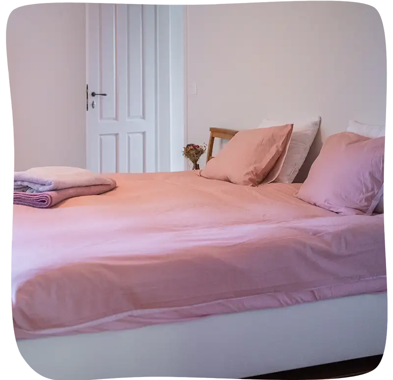 Lit  vu de côte et de très près avec des draps rose pale et un plaid sur la house de couette. On peut voir 3 coussins dont 2 rose pâle et deux blanc. Le sommier est blanc