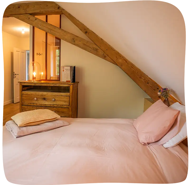 Lit avec des draps rose pâle et 2 coussins vu côté en premier plan avec une armoire en arrièrre plan et une charpente en bois surblombant le lit.