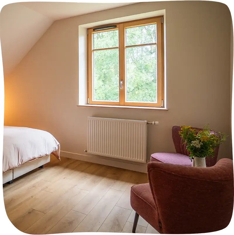 Une grande fenêtre lumineuse avec un radiateur en dessous au miliau, un bout du lit double aux draps rose pâle à gauche et deux fauteuils rouge à droite. On peut aperçvoir un pot de fleur entre les deux fauteuils.