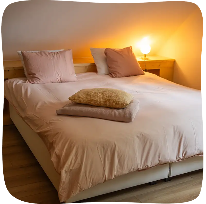 Lit aux draps rose pâle avec 4 coussins dont 2 blanc vu du bout et un petit peu de biais avec un plaid et un coussin supplémentaire dessus.