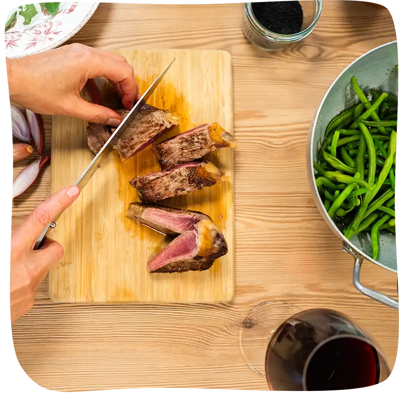 Couteau en train de découper une pièce de viande sur un comptoir avec des haricots dans une passoire en métal et un verre de vin sur le côté