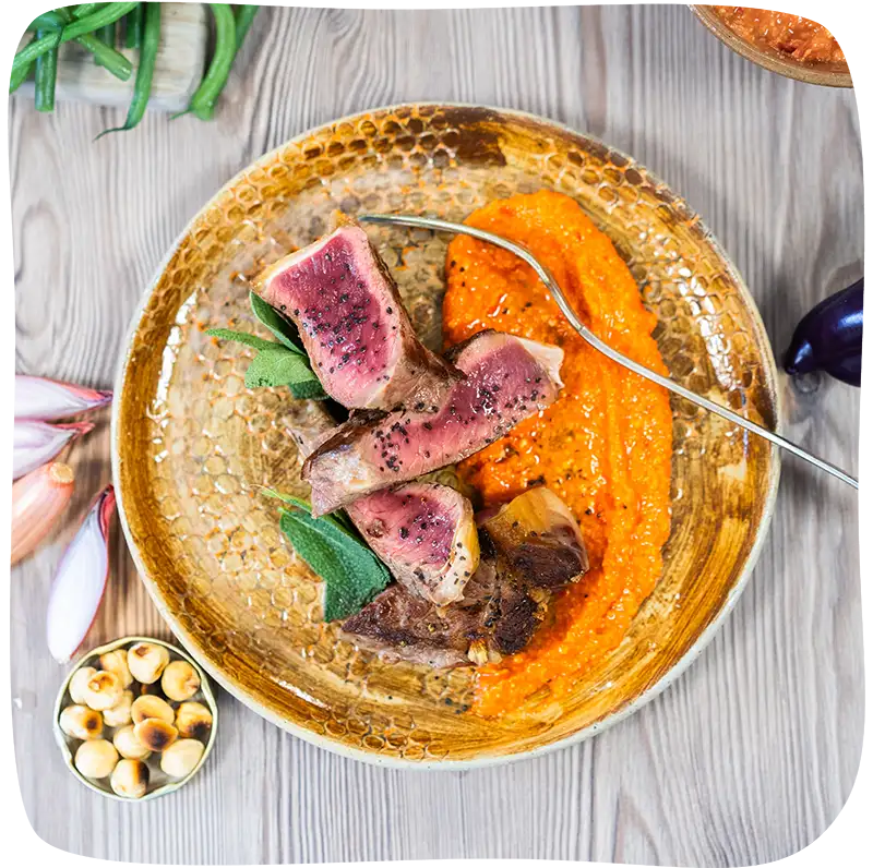 Assiette remplie de viande, une purée de carotte et d'une fourchette vue de haut sur une table en bois décorée d'échalotte et d'haricots.