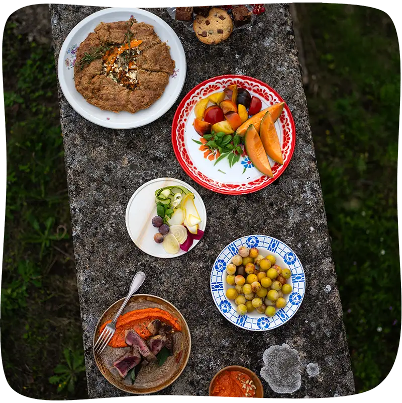 Table vue de haut composée de 5 assiettes en quinconce remplies et d'un présentoir en verre rempli de muffins et de cookies. L'assiette la plus en haut est remplie d'une galette bretonne, juste en dessous nous retrouvons une assiette de morceaux de melons et de fruits, suivie d'une assiette avec des tranches de légumes, suivie d'une assiette d'olives vertes et finalement une assiette remplie de viande, une purée de carotte et d'une fourchette.
