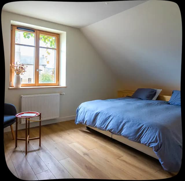 Chambre 1 - Lit double avec des draps bleus clairs. on voit la fenêtre sur le mur en face à gauche. On voit un petit tabouret rouge et un fauteul 1 place juste devant ce tabouret dans le coin à gauche