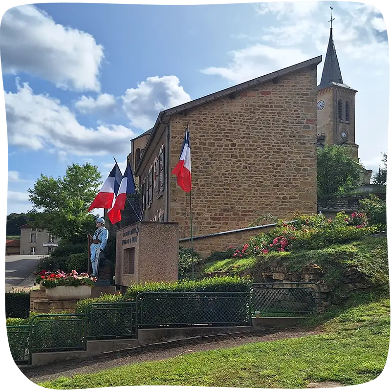Petite barrière verte avec une haie juste derrière en premier plan. Vue de côté d'un monument avec des drapeaux français au dessus et une statue d'un soldat devant en milieu de plan. On voit un maison en pierre de côté en arrière plan