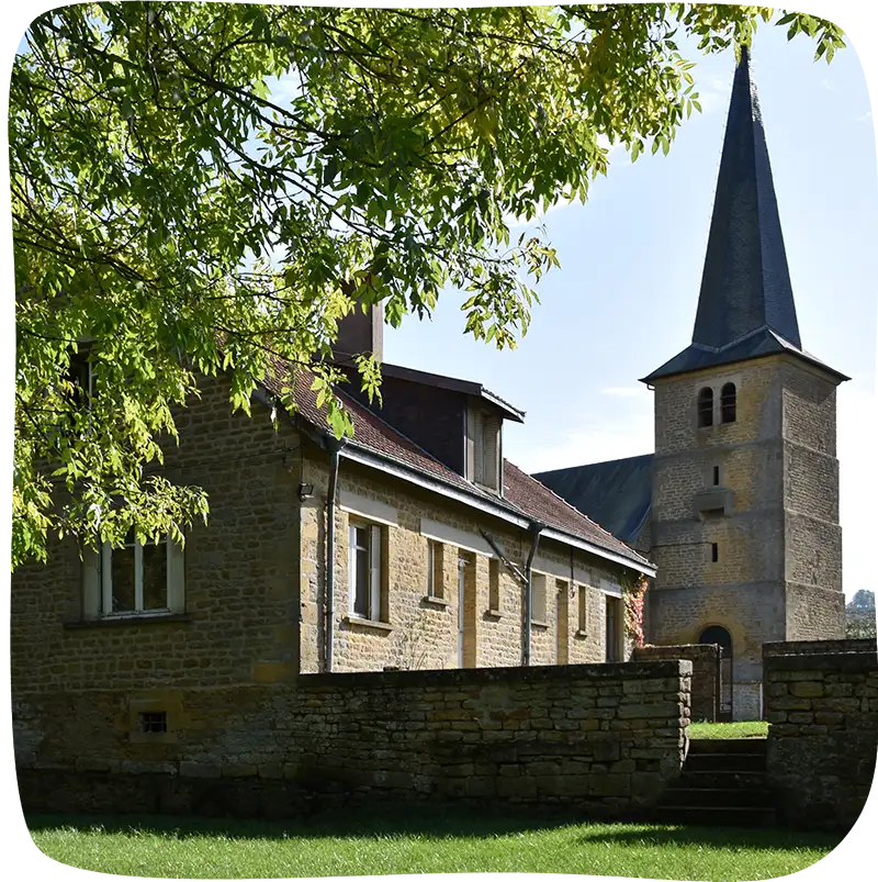 La maison d'hôte avec un muret devant et le soleil qui donne sur la façade. On retrouve l'église dans l'arrière plan.